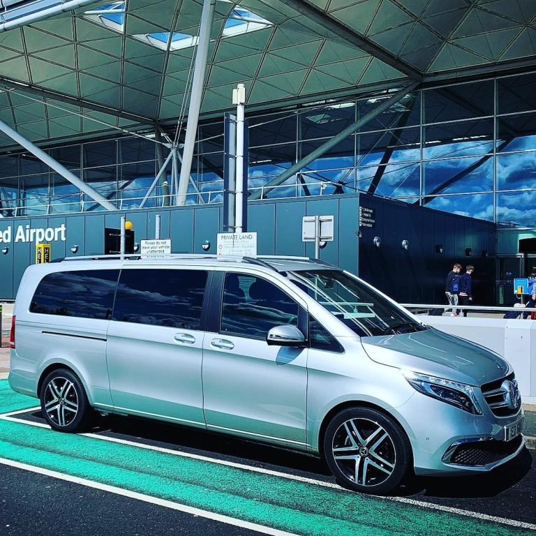 6 Seater Car in Airport A silver six-seater car is parked at an airport, with an airport building in the background.